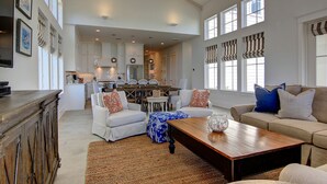 A spacious living area with a kitchen in the background, featuring a high ceiling, large windows, and a mix of modern and classic furniture.