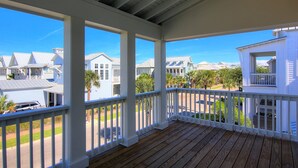 A covered porch with white railings and wooden decking, overlooking a residential area with houses and palm trees.