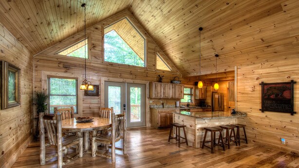 A rustic cabin interior with a wooden dining table, chairs, a kitchen area, and a large window.