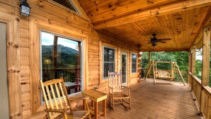 A wooden cabin with a porch, two rocking chairs, a table, and a ceiling fan.