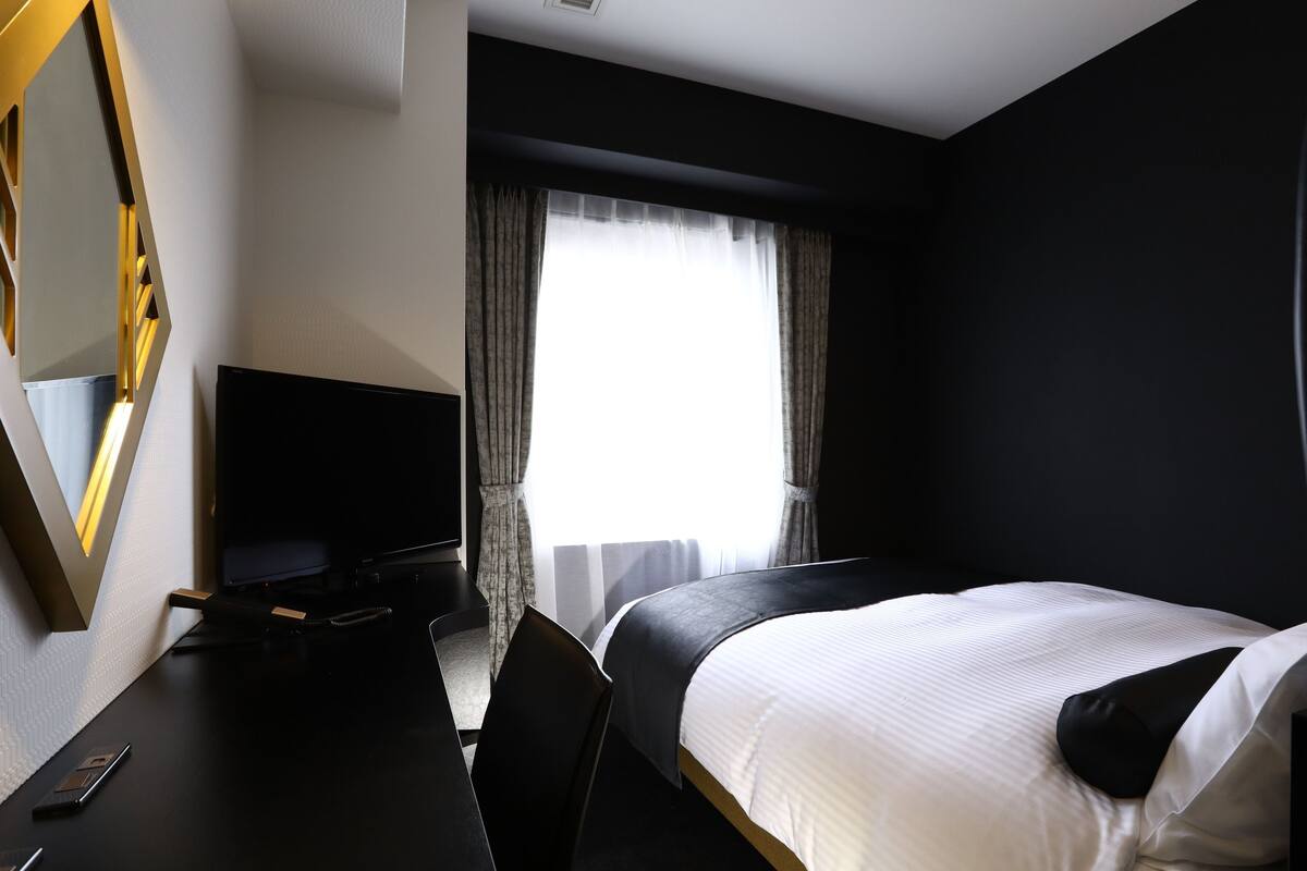 Une chambre d’hôtel avec un lit, un bureau avec une télévision, une chaise et une fenêtre avec des rideaux.