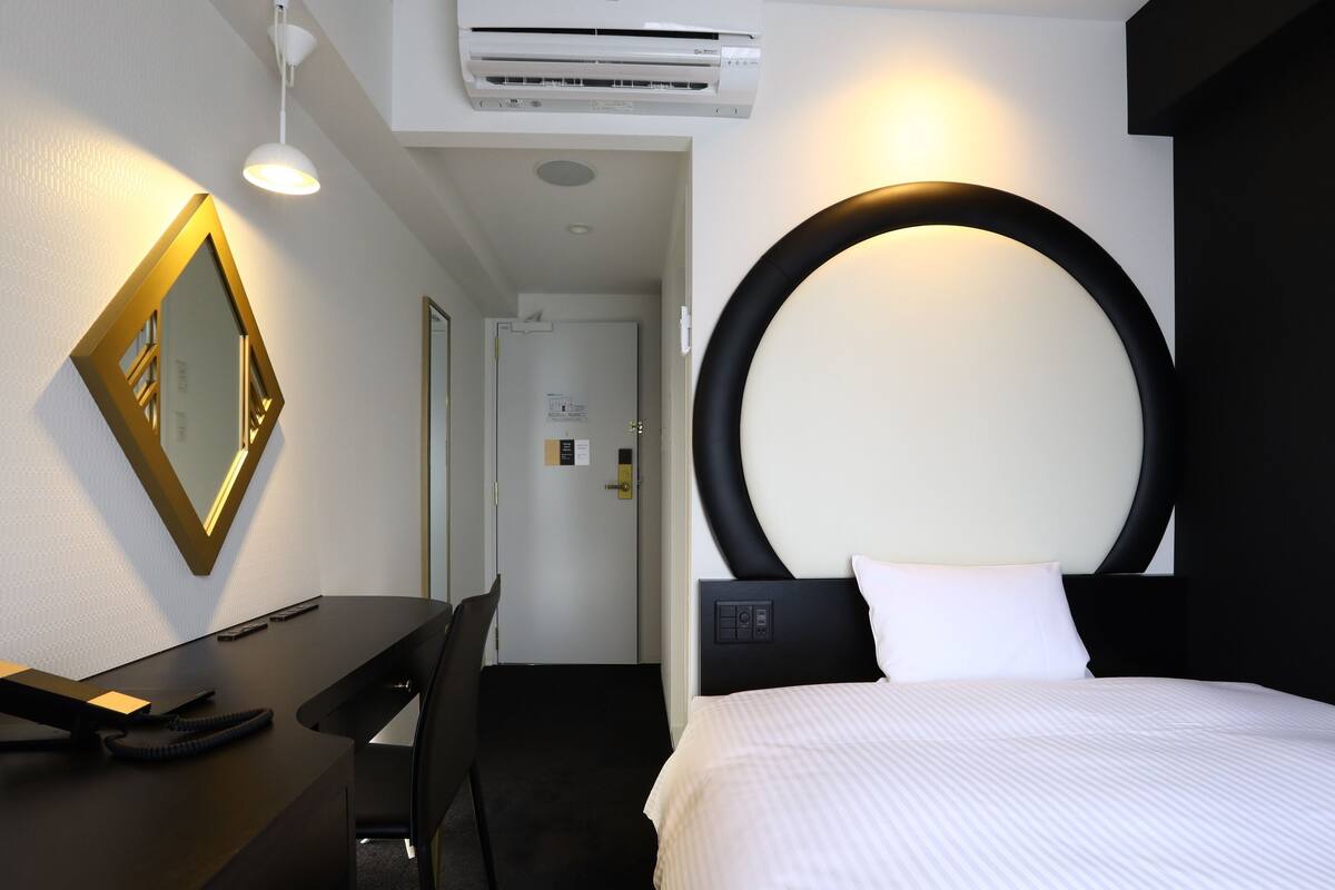 Une chambre d’hôtel équipée d’un lit, d’un bureau, d’une chaise, d’un luminaire rond fixé au mur et d’un appareil de climatisation.