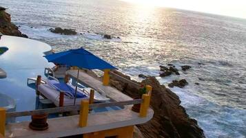 Uma piscina infinita na encosta de um penhasco, com uma espreguiçadeira e guarda-sol, com vista para o oceano.