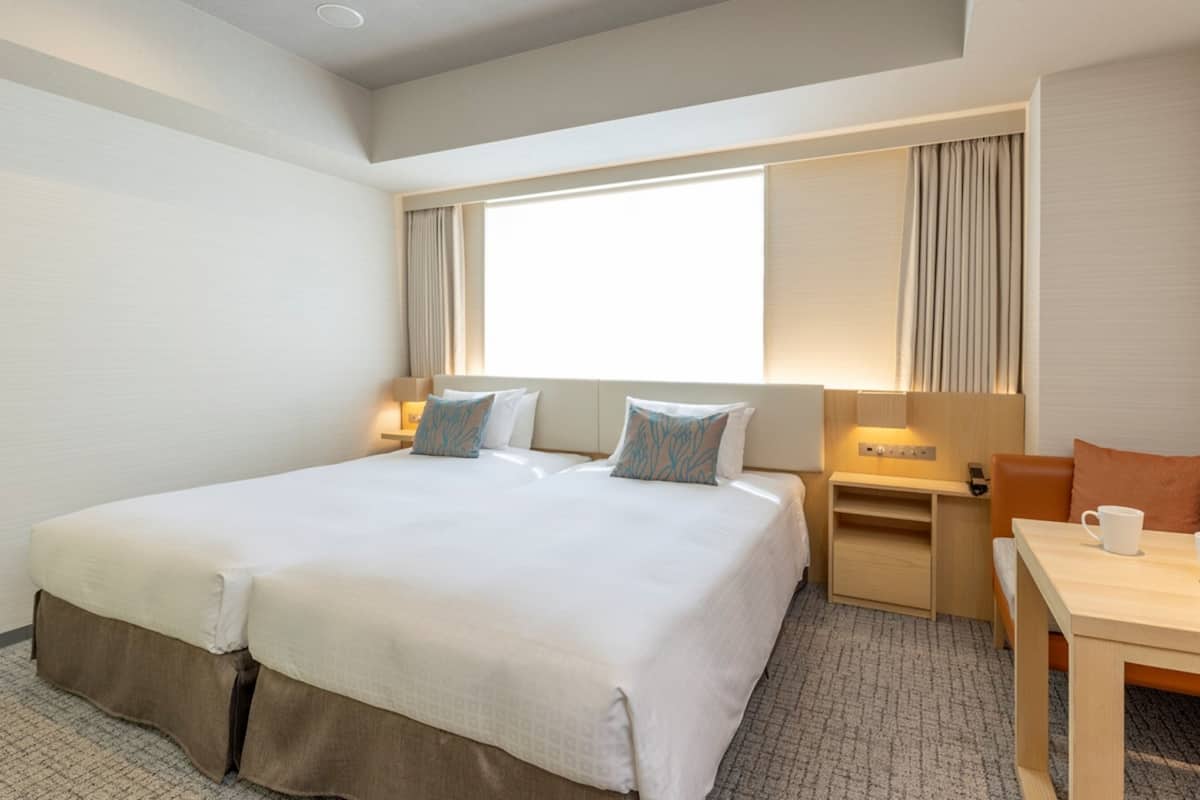 Une chambre d’hôtel avec deux lits, un bureau, une chaise, une petite table et une fenêtre avec des rideaux.