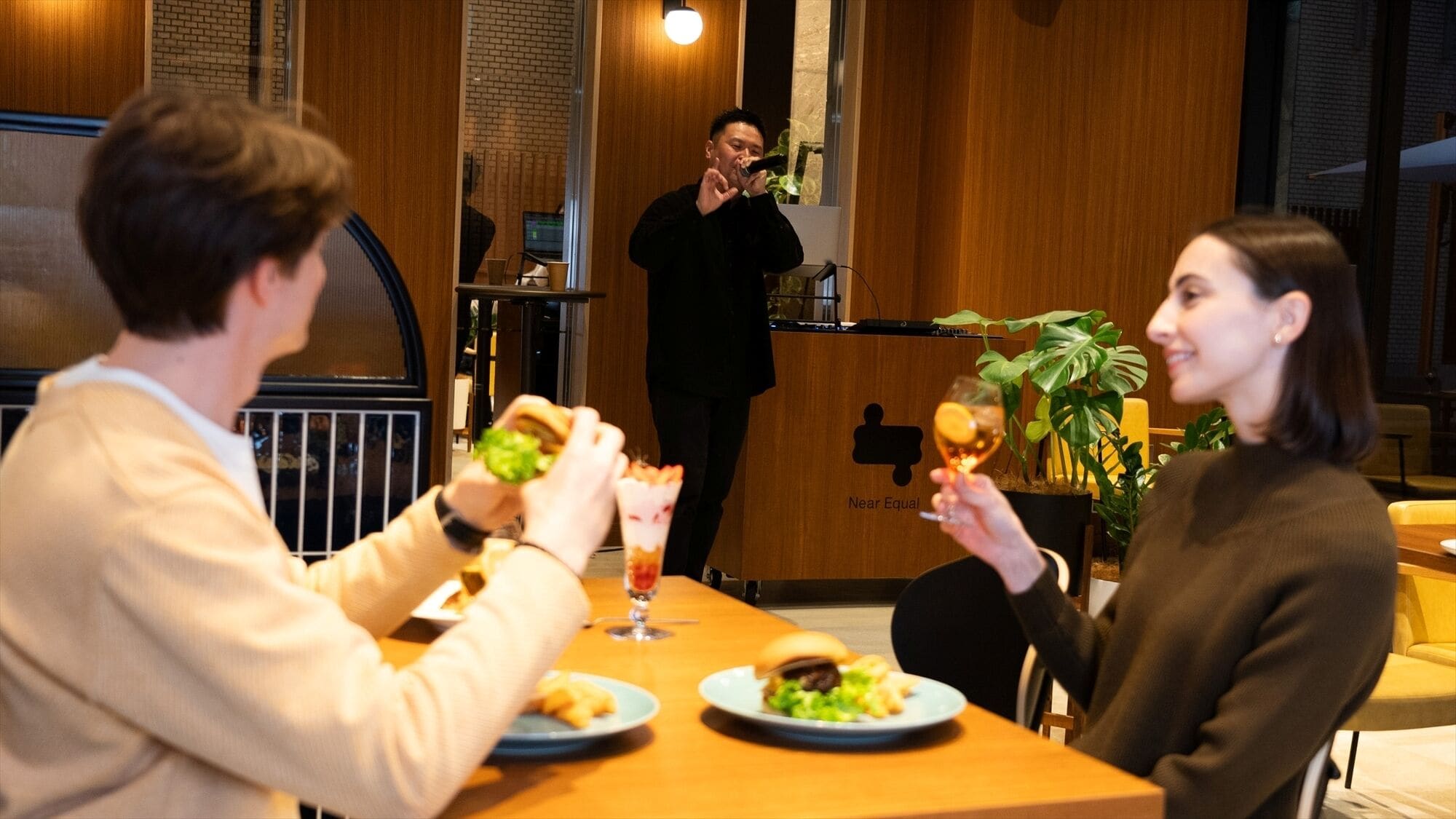 Ambiente de restaurante com paredes de madeira e decoração moderna. Duas pessoas jantam à mesa, com pratos de comida e bebidas. Uma pessoa está ao fundo, possivelmente um garçom ou funcionário.