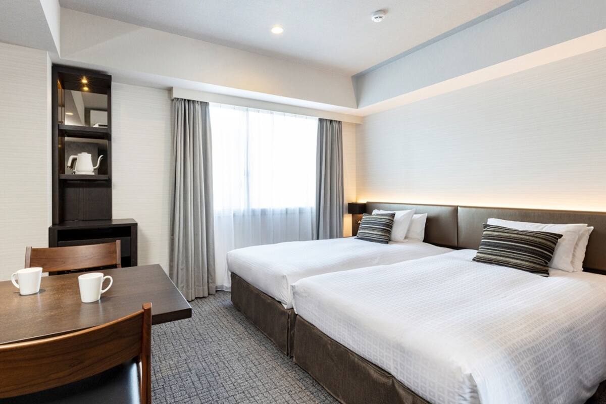 Une chambre d’hôtel avec deux lits, un bureau avec une chaise, une petite étagère avec une théière et une fenêtre avec des rideaux.
