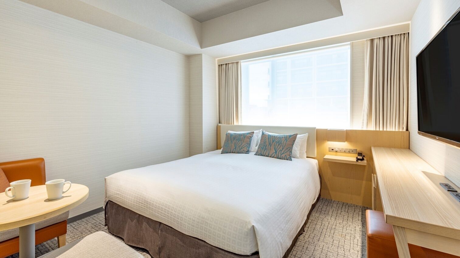 A hotel room with a bed, a desk, a chair, a small table, and a TV.