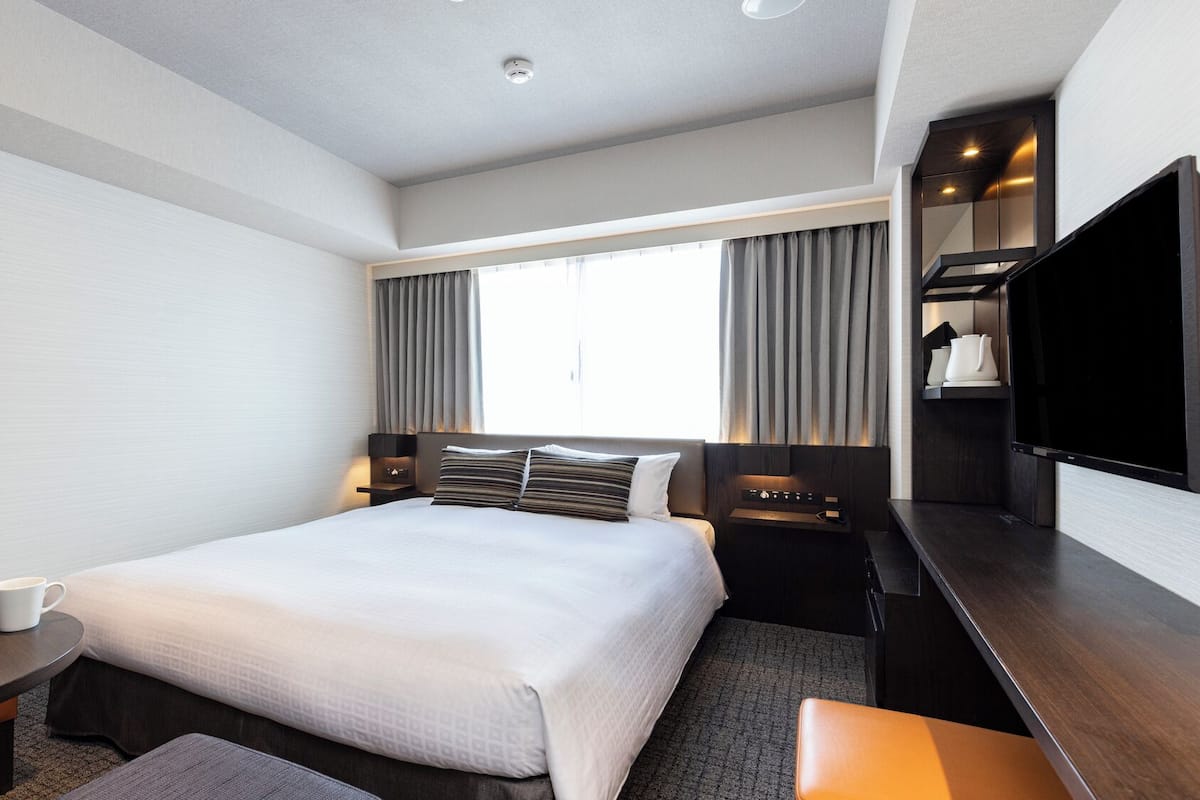 Une chambre d’hôtel avec un lit, un bureau, une chaise, une télévision et une fenêtre avec des rideaux.