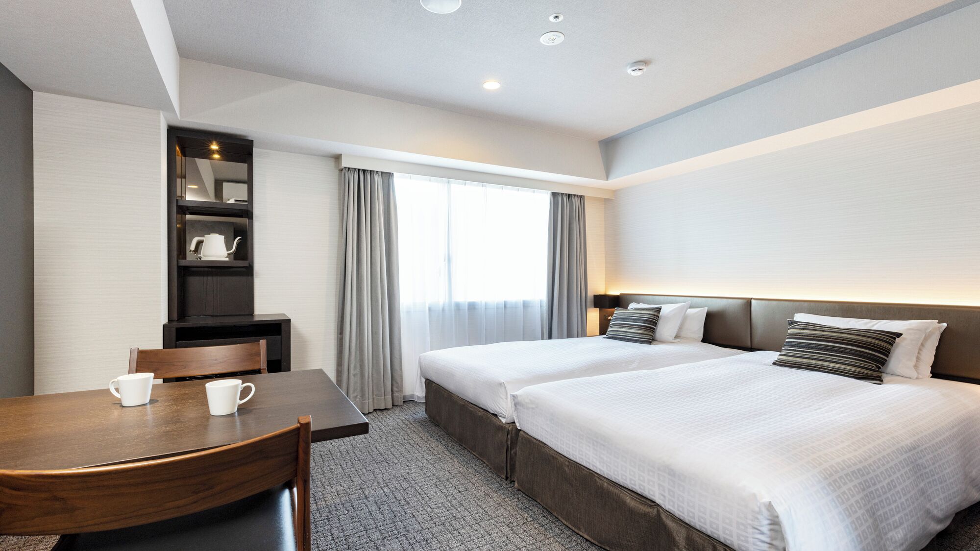 A hotel room with two beds, a desk, a chair, a bookshelf, and a window with curtains.