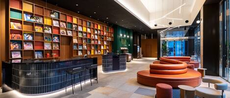 Un moderno vestíbulo de hotel con un mostrador de recepción central, una librería colorida y una zona de estar con sillas naranja y grises.