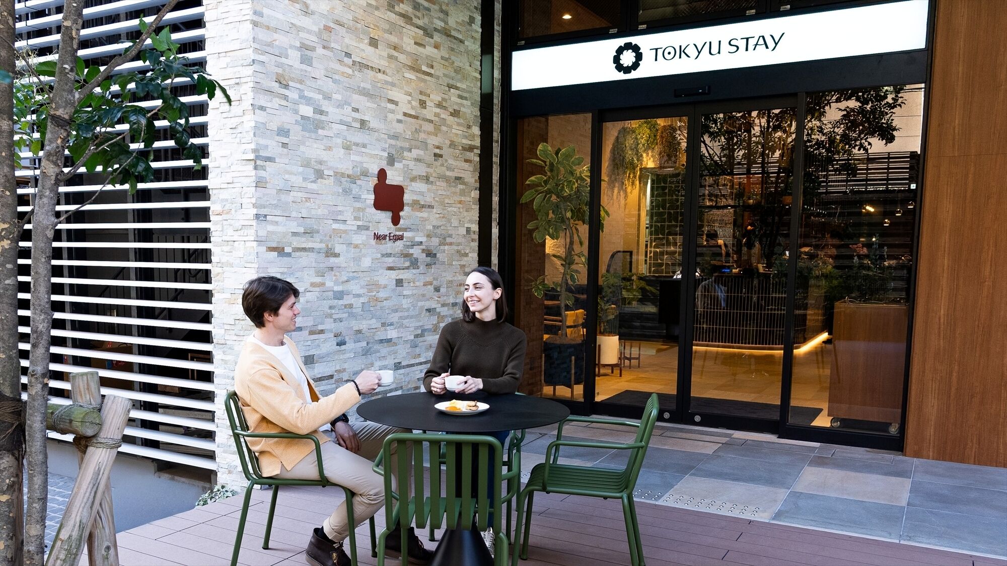 Deux personnes sont assises à une table à l’extérieur, et on peut voir en arrière-plan une enseigne indiquant « TOKYU STAY ».
