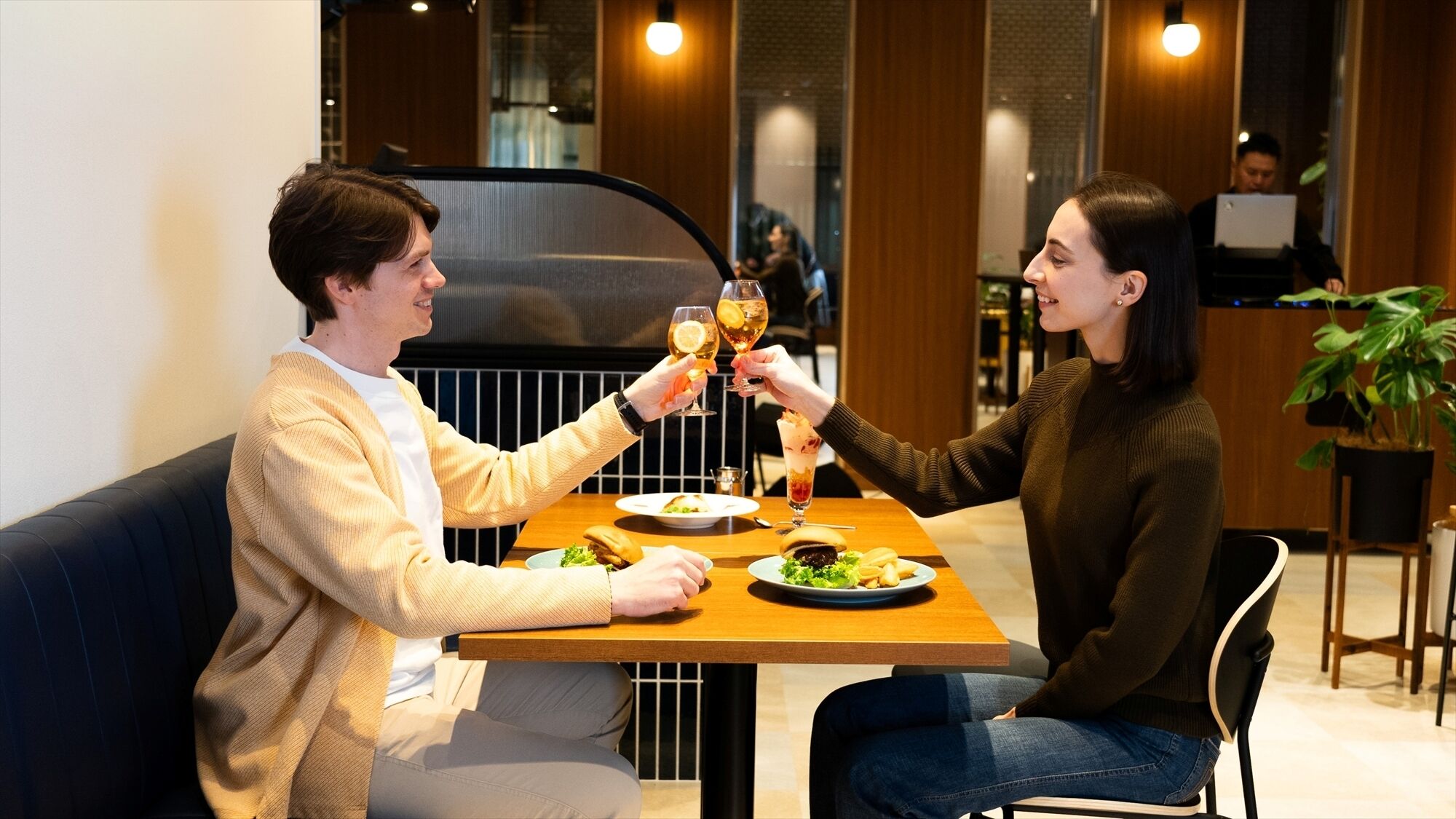 Twee personen proosten aan een tafel met eten en drinken, in een moderne, overdekte setting met houten details en een plant.