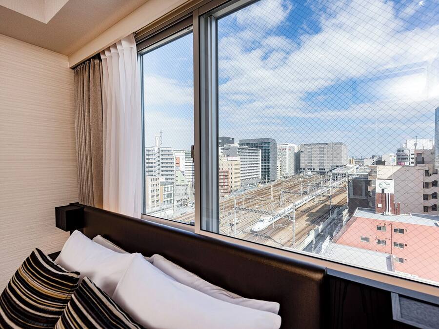 A hotel room with a large window offering a city view, a bed with striped pillows, and a nightstand with a lamp.