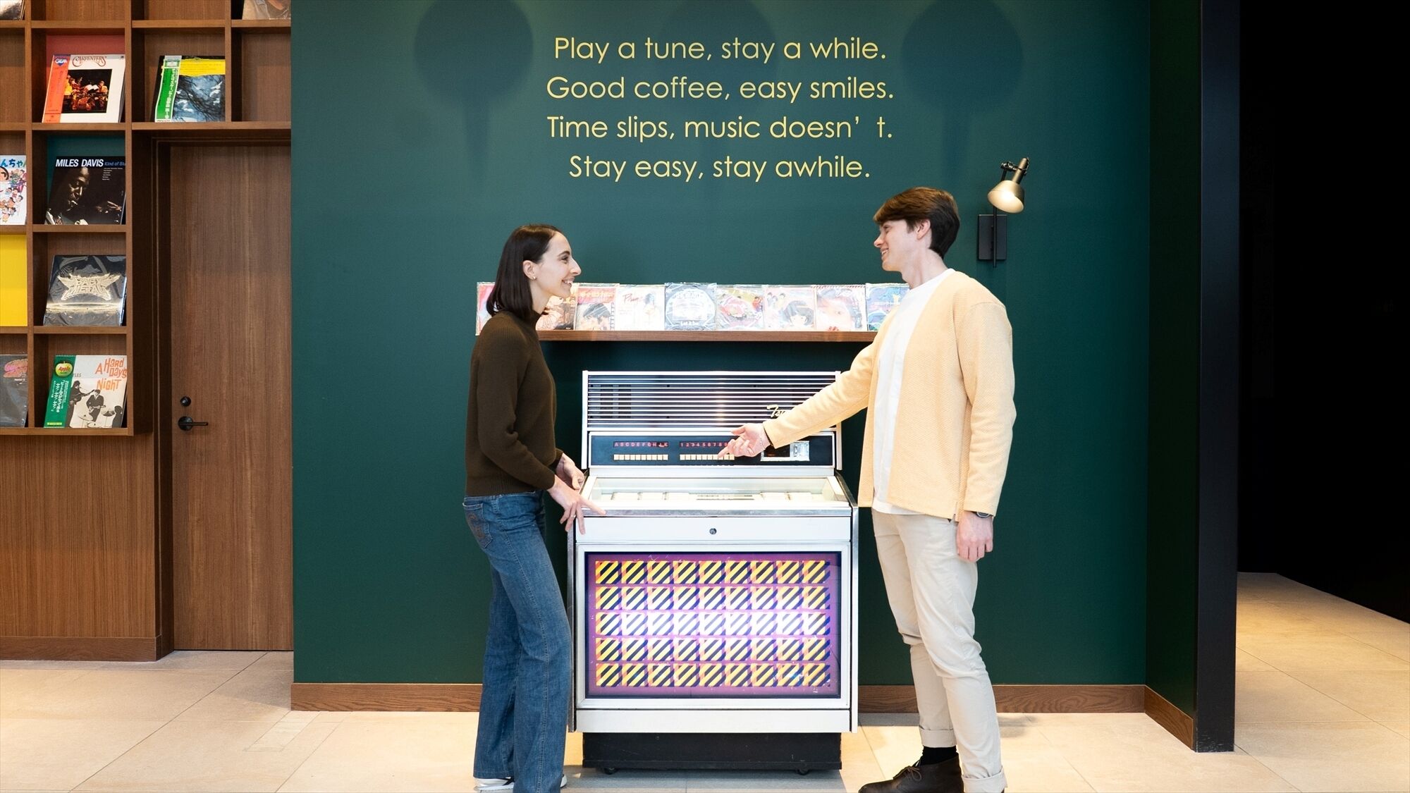 A man and a woman interacting with a jukebox in a room with a green wall and a bookshelf filled with records.