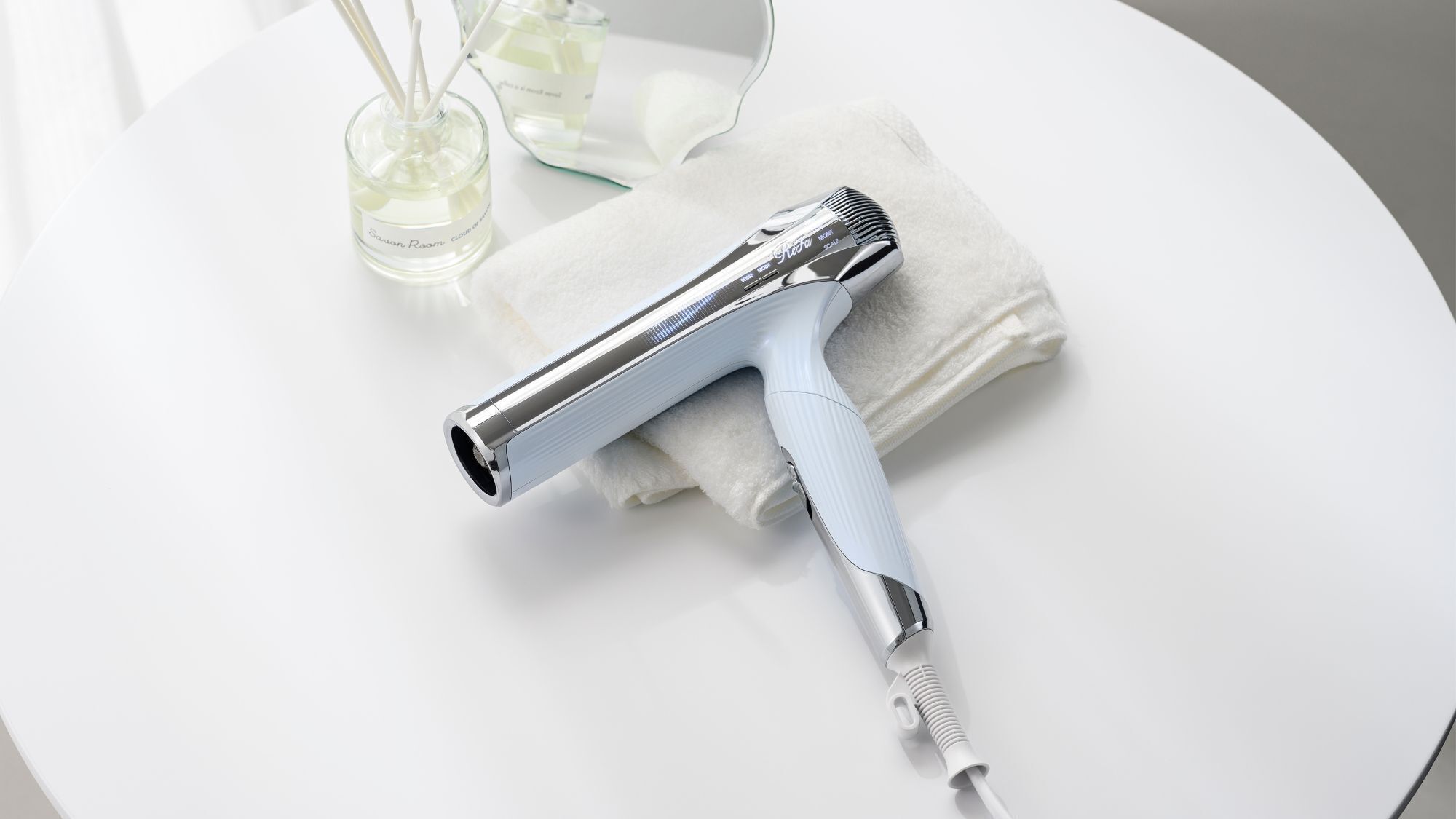 A hair dryer on a white surface with a towel and a glass container with a liquid.
