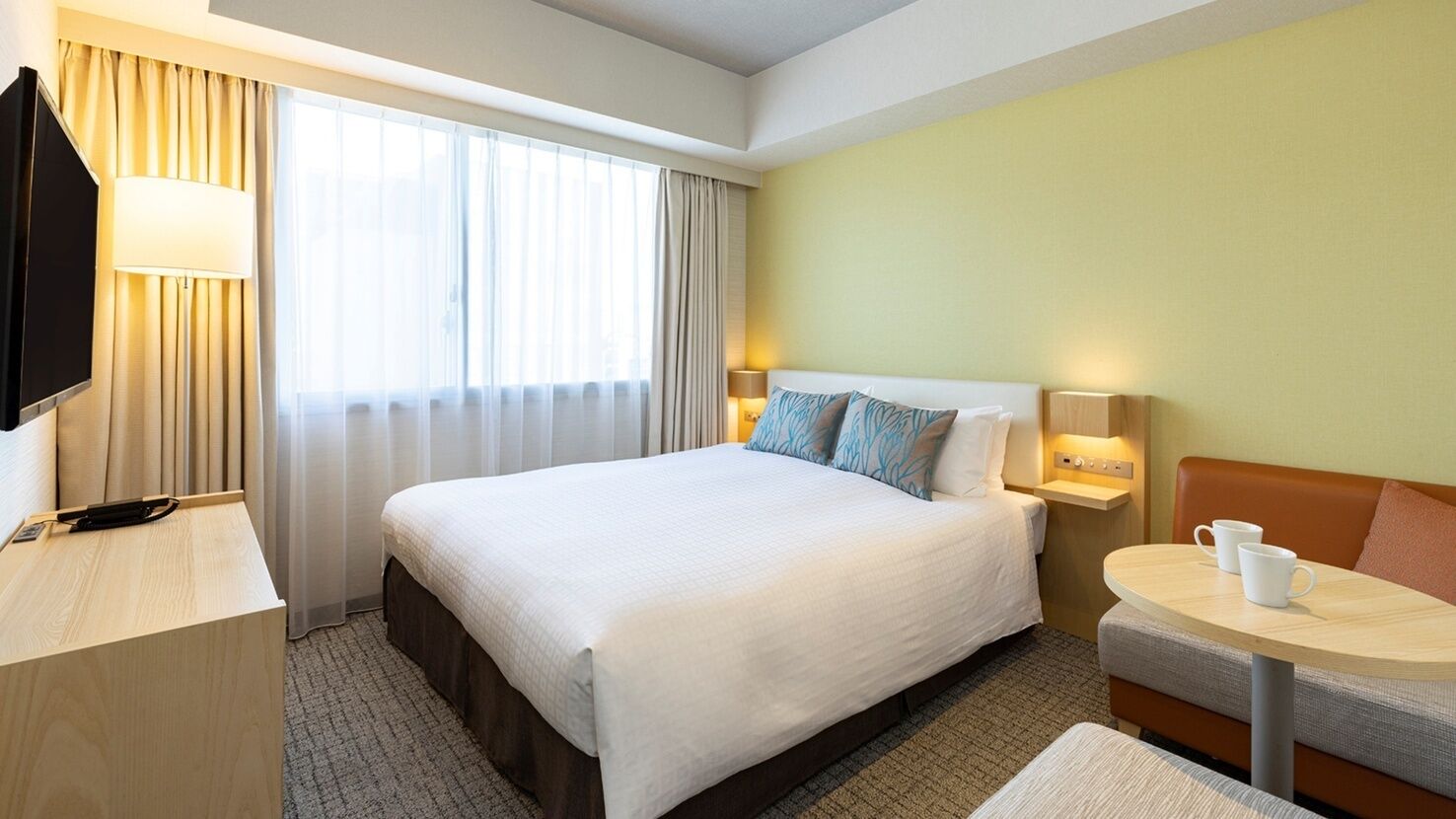 A hotel room with a bed, bedside tables, a TV, a desk, and a small table with a cup.