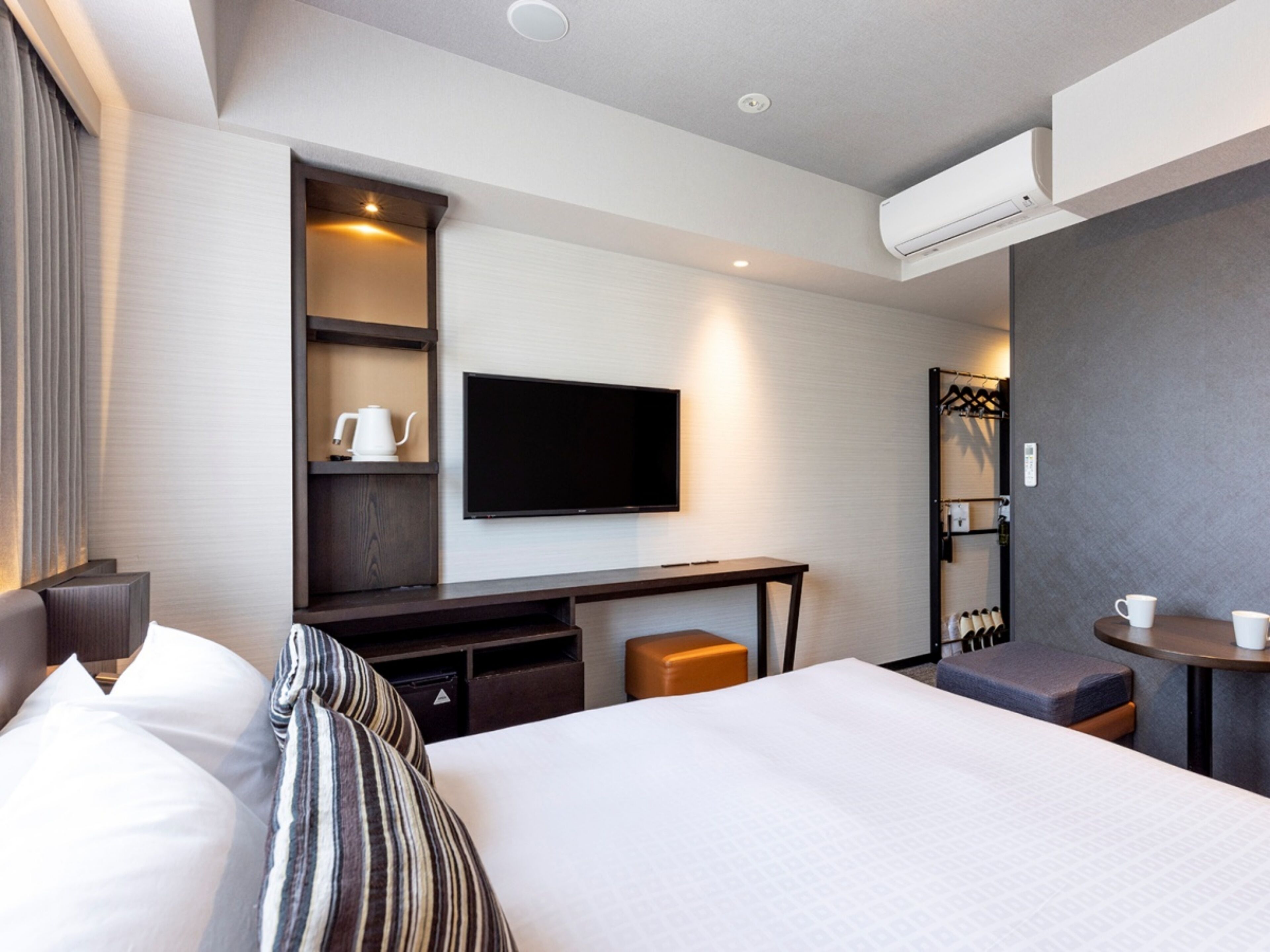 A modern hotel room with a large bed, a flat-screen TV mounted on the wall, a desk with a chair, a small table with a cup, and a shoe rack.