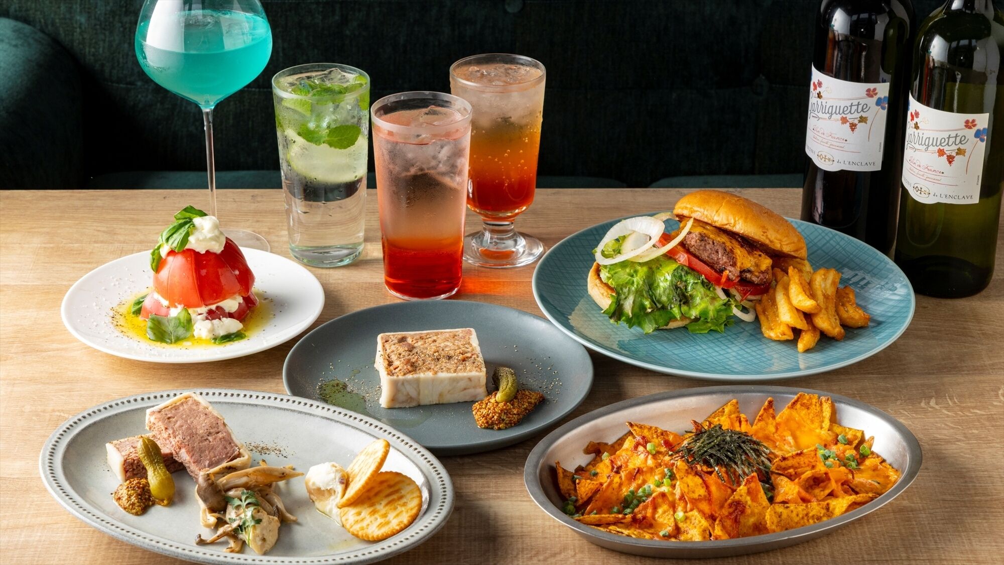 Variedad de platos que incluyen una hamburguesa, papas fritas, nachos y una ensalada, junto con dos vasos de una bebida roja y una botella de vino.