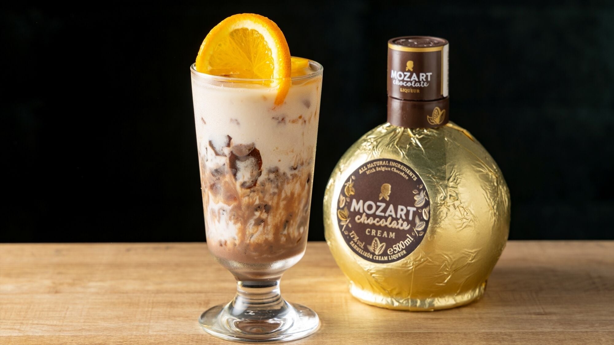 A glass of chocolate liqueur with an orange slice garnish and a bottle of Mozart chocolate liqueur beside it.