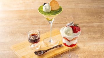 A dessert tray with two desserts on a wooden board, a spoon, and a small glass of syrup.