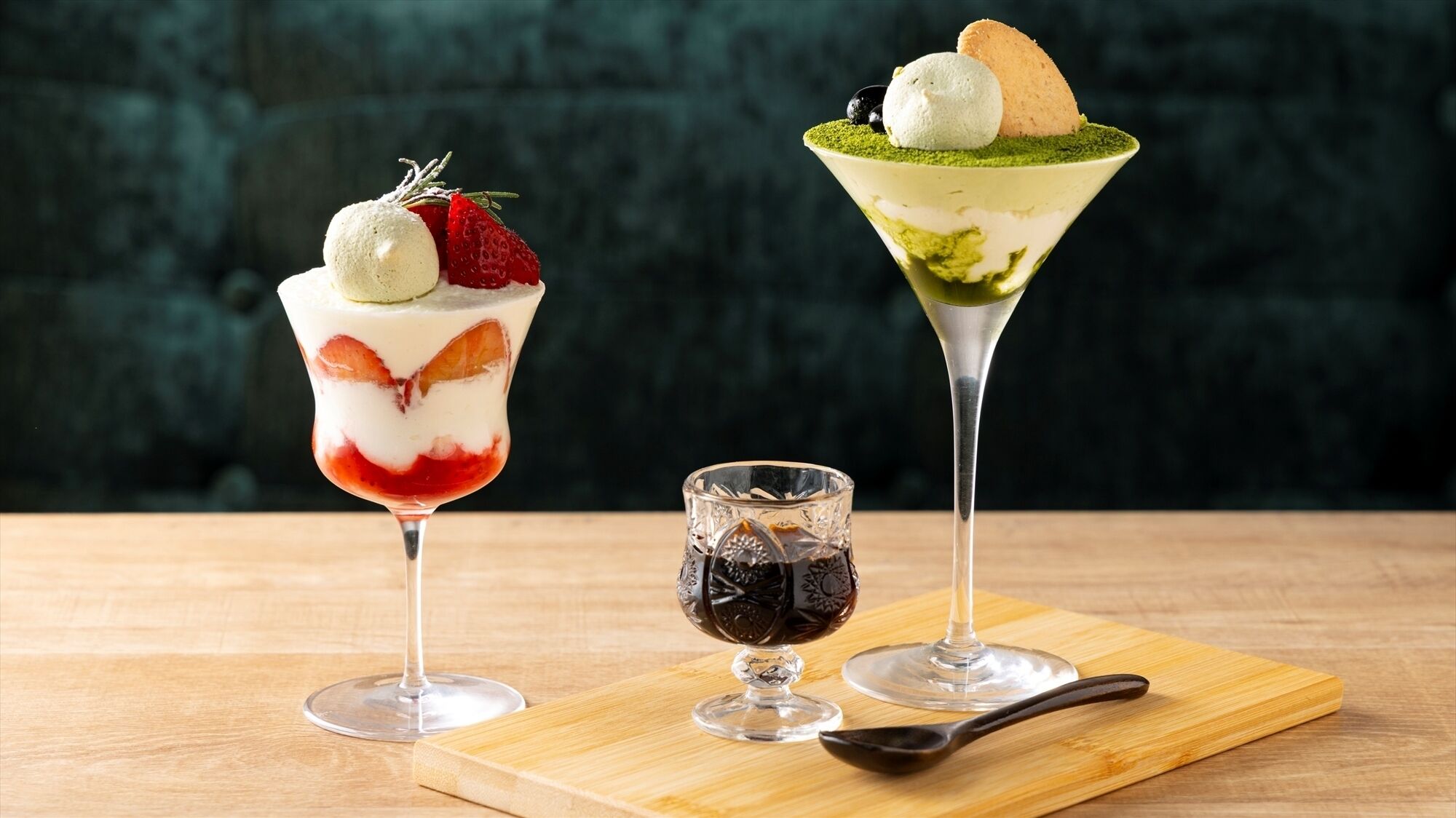 Dos vasos de postre sobre una tabla de madera con una cuchara, y un vaso pequeño de líquido oscuro a un lado.