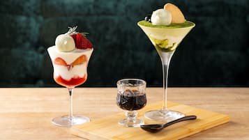 Dos vasos de postre sobre una tabla de madera con una cuchara, y un vaso pequeño de líquido oscuro a un lado.