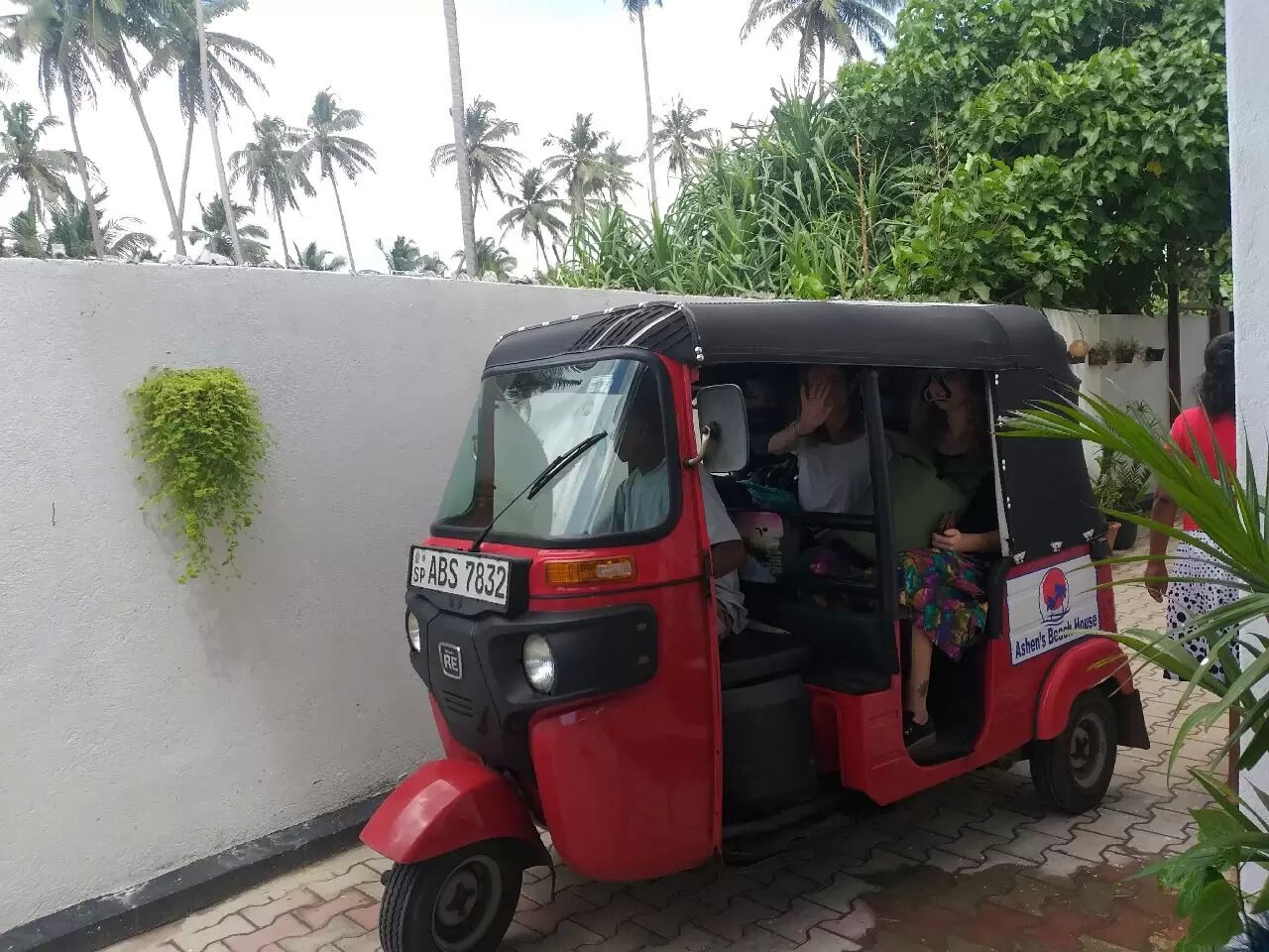 Un tuk-tuk rojo estacionado en una zona pavimentada, con un muro blanco y plantas verdes al fondo.