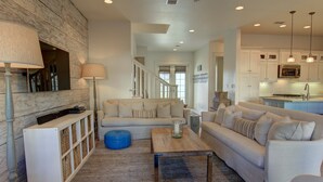 A modern living room with a sofa, coffee table, and ottoman, a wooden accent wall, and a kitchen area with cabinets and a microwave. - Lakefront Beach Home@Cinnamon Shore North w/ Sunset Views+Amenity Access! (Port Aransas)