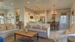 A spacious living room with a sofa, coffee table, and ottoman, a dining area with chairs, and a kitchen with modern appliances and pendant lighting. - Lakefront Beach Home@Cinnamon Shore North w/ Sunset Views+Amenity Access! (Port Aransas)