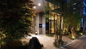 Una entrada de hotel moderna con un camino de piedra, iluminada por luces en el suelo, rodeada de árboles y una fuente pequeña.