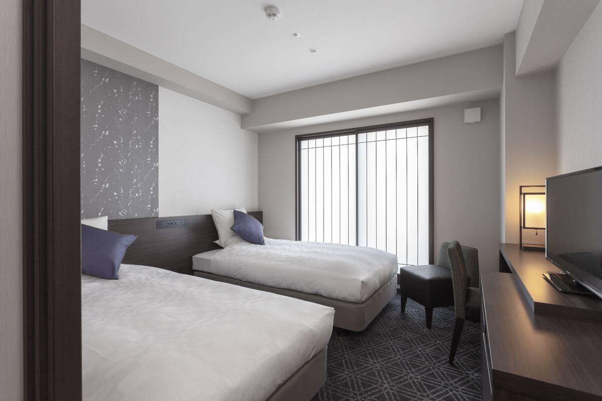 A hotel room with two beds, a desk, a chair, a TV, and a large window.