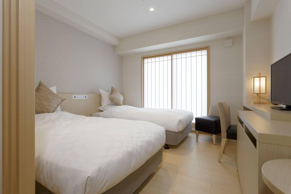 A hotel room with two beds, a desk, a chair, and a television.
