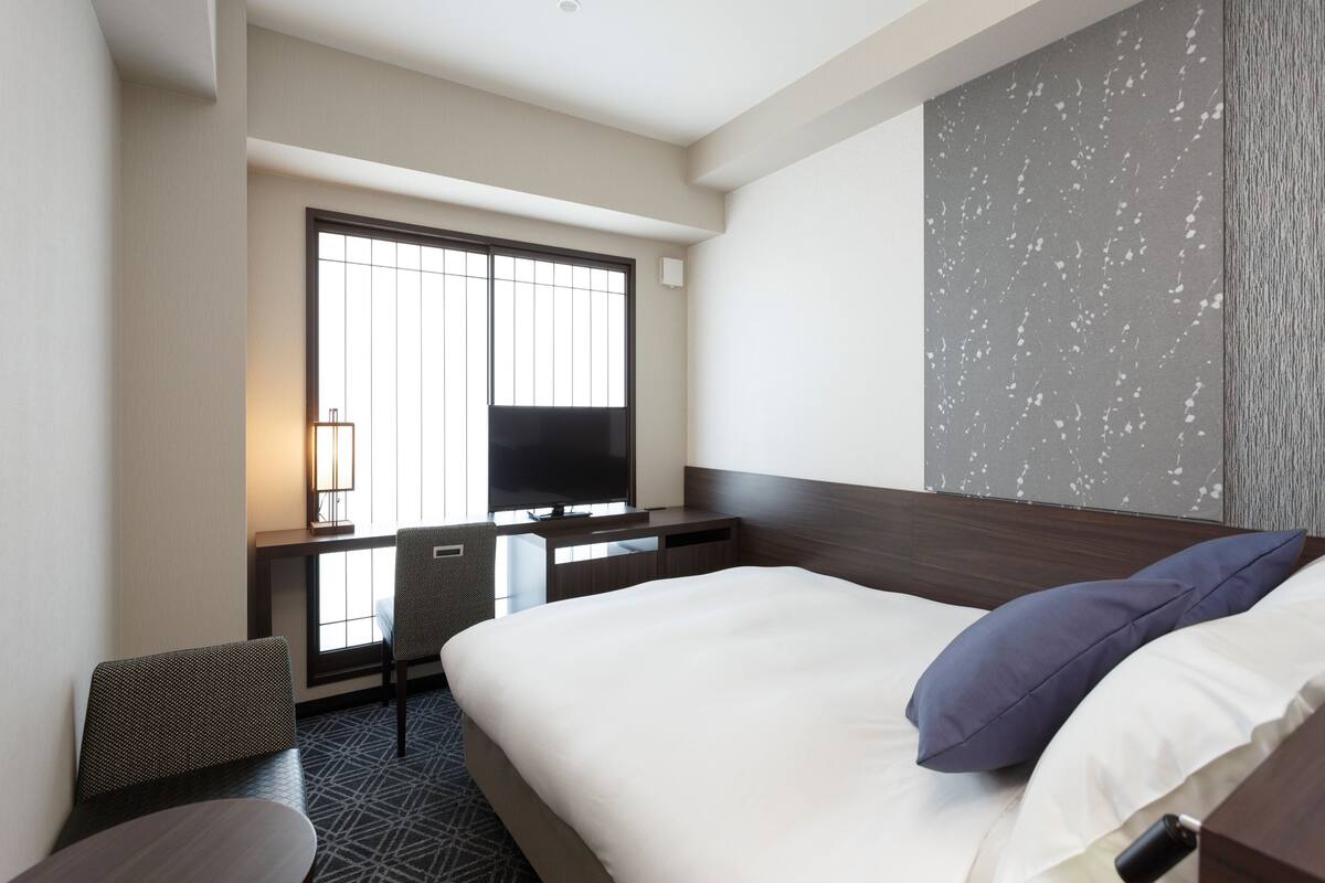 A hotel room with a bed, a desk with a chair, a TV, and a window.