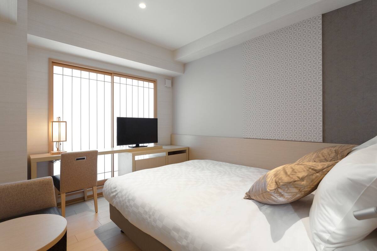 A hotel room with a bed, a desk with a TV, a chair, and a window.