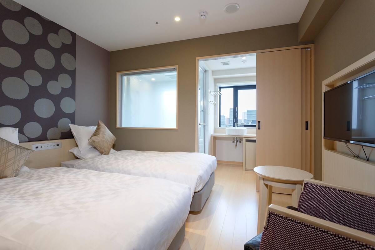 A hotel room with two beds, a small table, a TV, and a view of the city through a window.