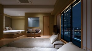 Habitación de hotel con una cama grande, una cama más pequeña, una zona de estar con una silla, un televisor y vistas a un paisaje urbano nocturno.