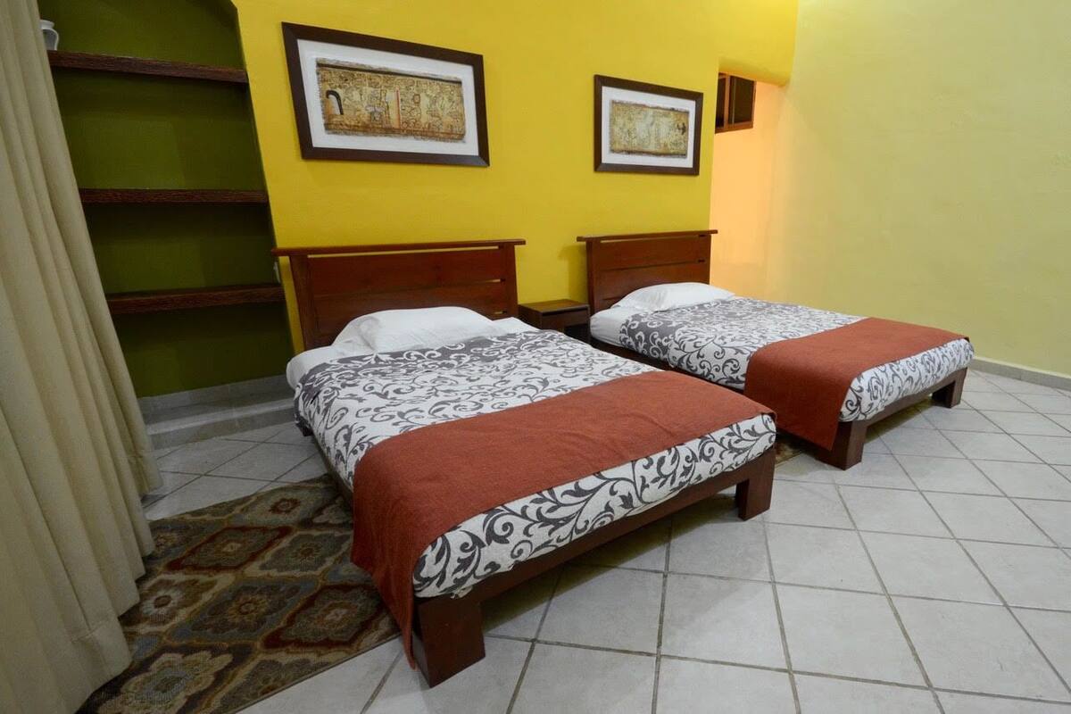 Una habitación con dos camas, cada una con una colcha estampada y una manta roja. Hay cuadros enmarcados en la pared y una alfombra estampada en el suelo.