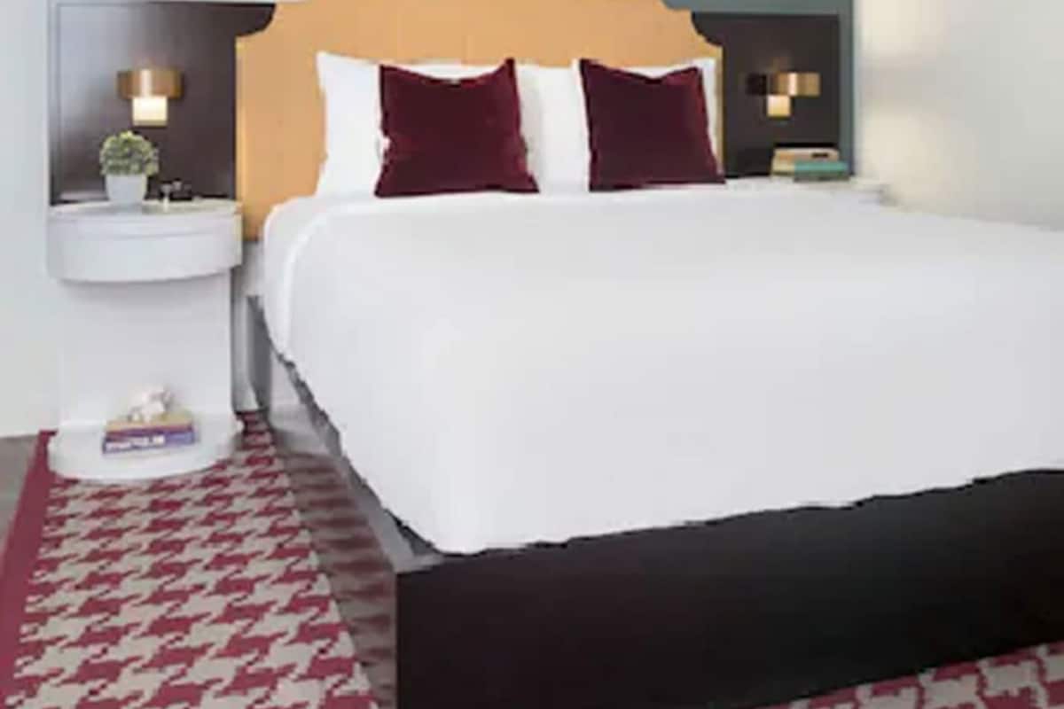 Une chambre d’hôtel avec un lit, une table de chevet, une commode, un tapis à motifs rouges et blancs, et un mur orné de rayures verticales.