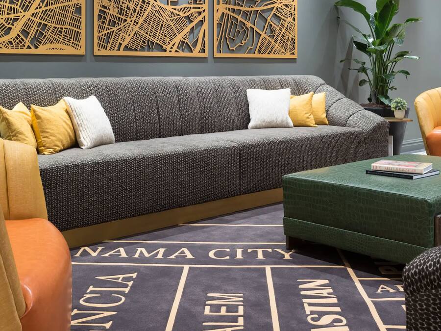 Una sala de estar moderna con un sofá gris, cojines amarillos y blancos, un otoman verde y una alfombra con motivos urbanos.