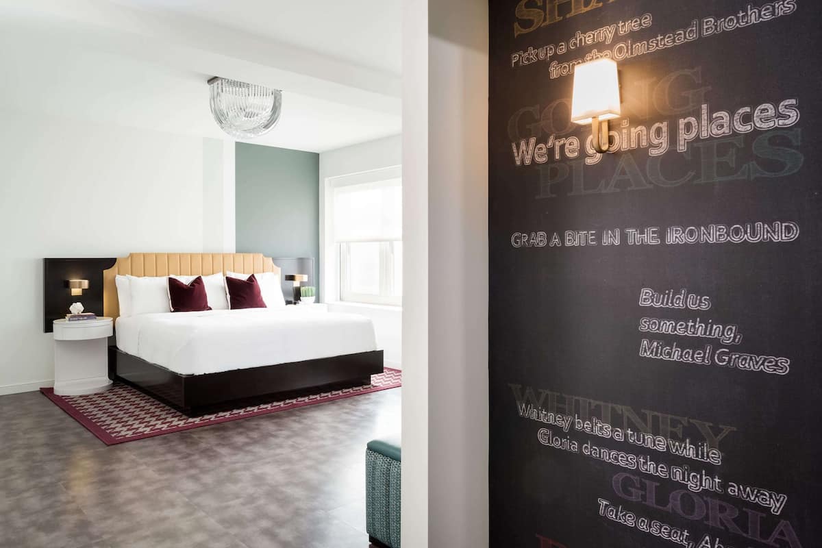 Une chambre d’hôtel moderne avec un grand lit, une table de chevet, une commode, un lustre et un mur orné d’un texte.