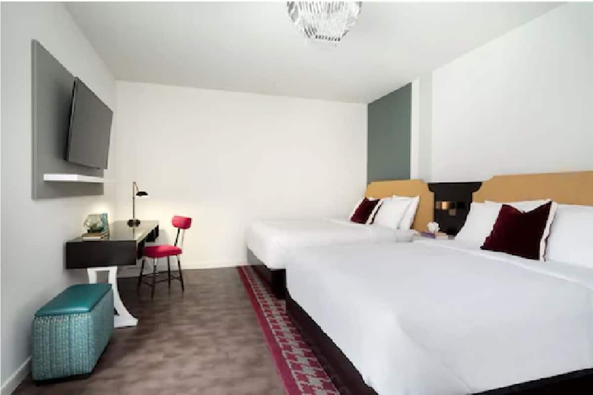 Une chambre d’hôtel avec deux lits, un bureau avec une chaise, un téléviseur fixé au mur et un petit pouf.