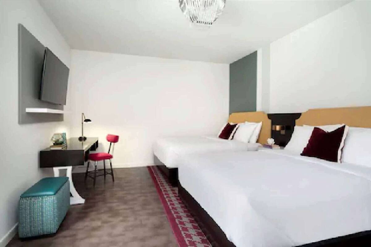 Une chambre d’hôtel avec deux lits, un bureau avec une chaise, un téléviseur fixé au mur et un petit tapis rouge.