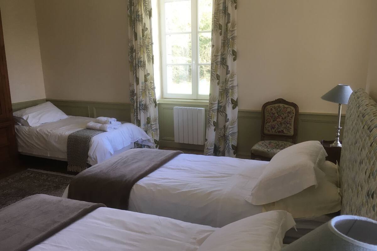 Una habitación con dos camas individuales, una silla, una mesa con lámpara y una ventana con cortinas.