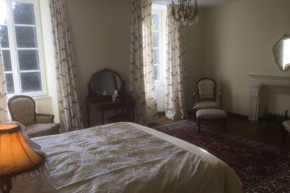Un dormitorio con cama, dos sillas, un tocador con espejo, una araña y una chimenea.
