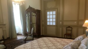 A bedroom with a large bed, a wooden wardrobe, a chair, a window, and a chandelier.