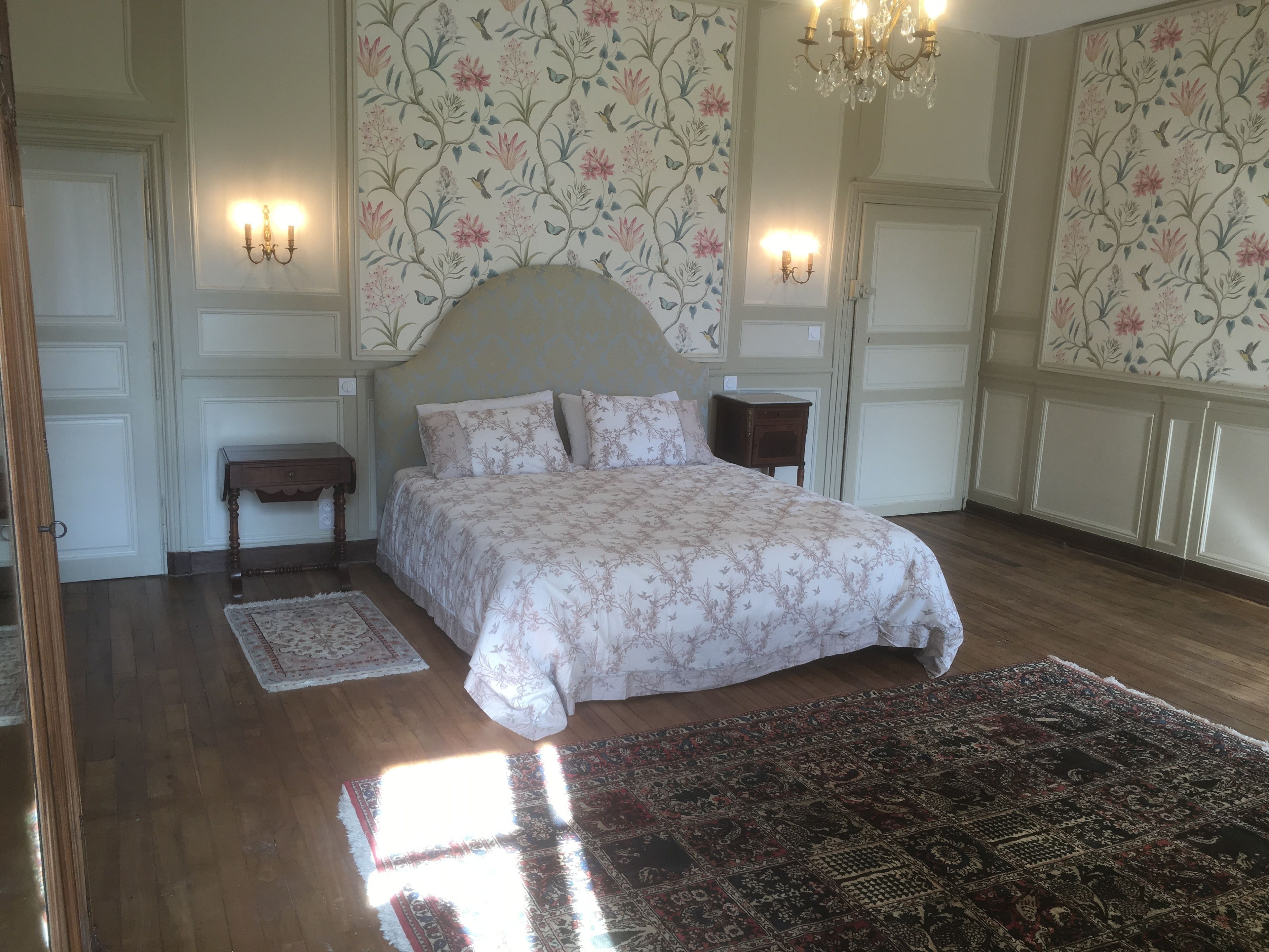 Un dormitorio con una cama grande, dos mesitas de noche, una araña, papel tapiz floral y una alfombra estampada.