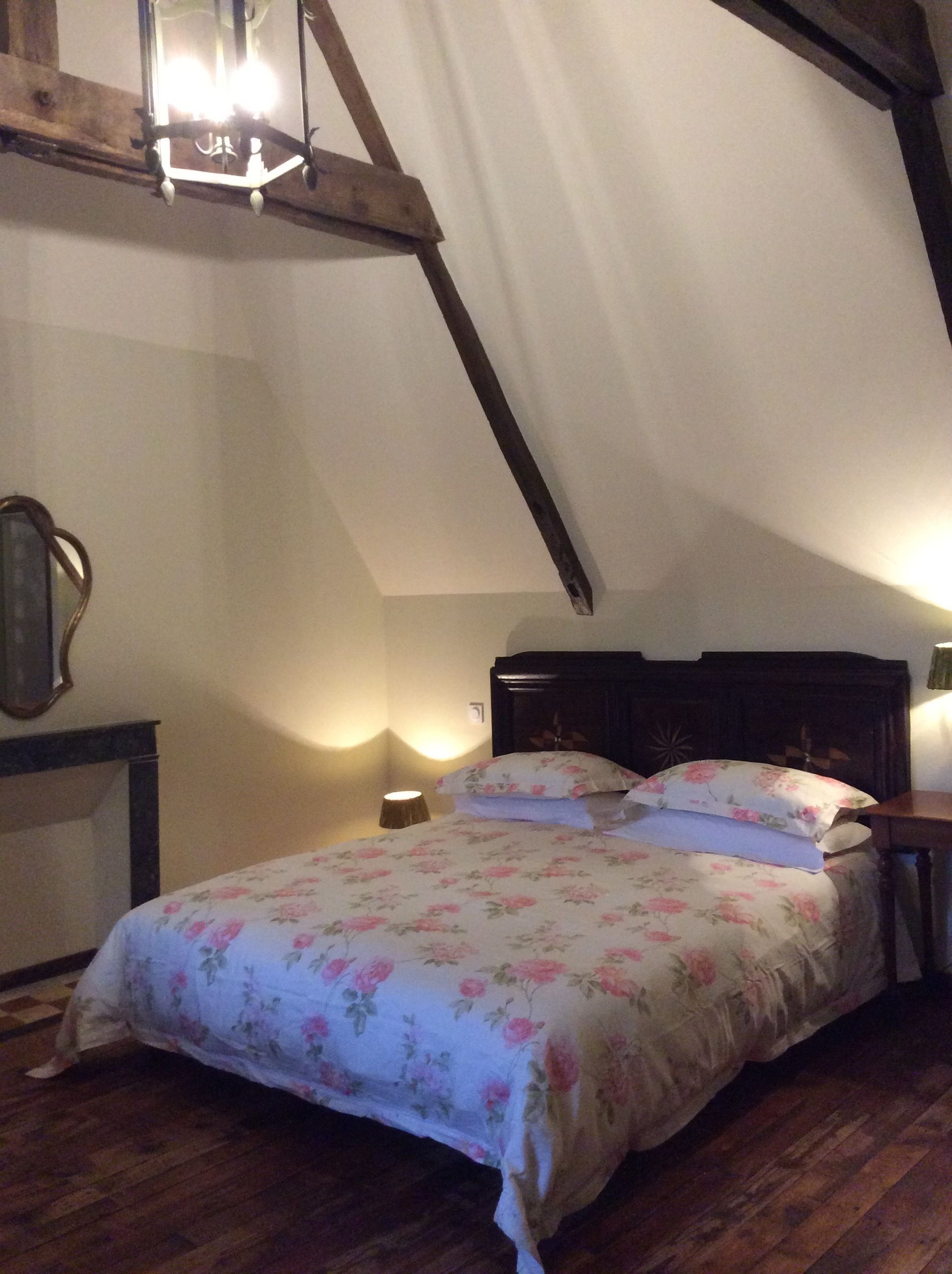 A bedroom with a sloped ceiling, wooden beams, a bed with floral bedding, a nightstand, and a wall-mounted mirror.