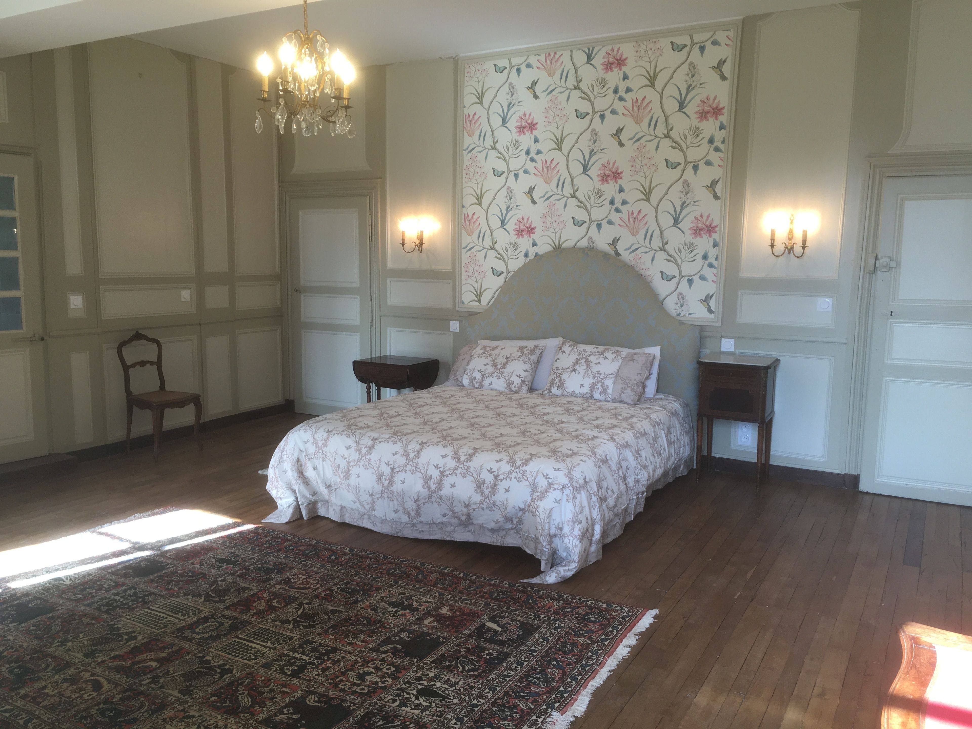 A bedroom with a large bed, a chandelier, a floral wallpaper, and a patterned rug.