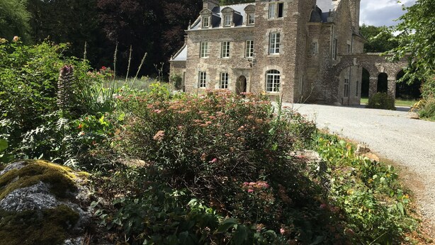 A historic stone building with a prominent tower, surrounded by lush gardens and a gravel driveway.