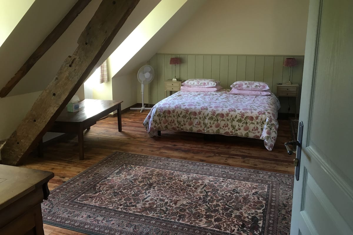 Un dormitorio con techo inclinado, piso de madera, cama con ropa de cama floral, una mesita de noche y una alfombra estampada.
